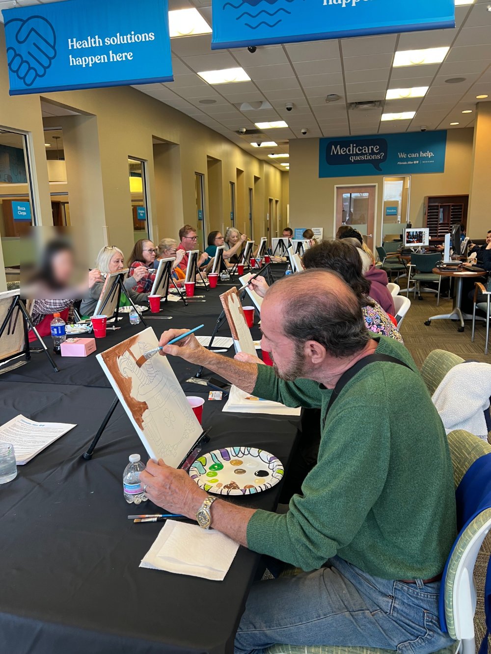 A group of people participating in a painting class at a community center or similar venue. They are seated at a long table with individual canvases, some of which are decorated or sketched, and are painting with brushes. The room has signs related t