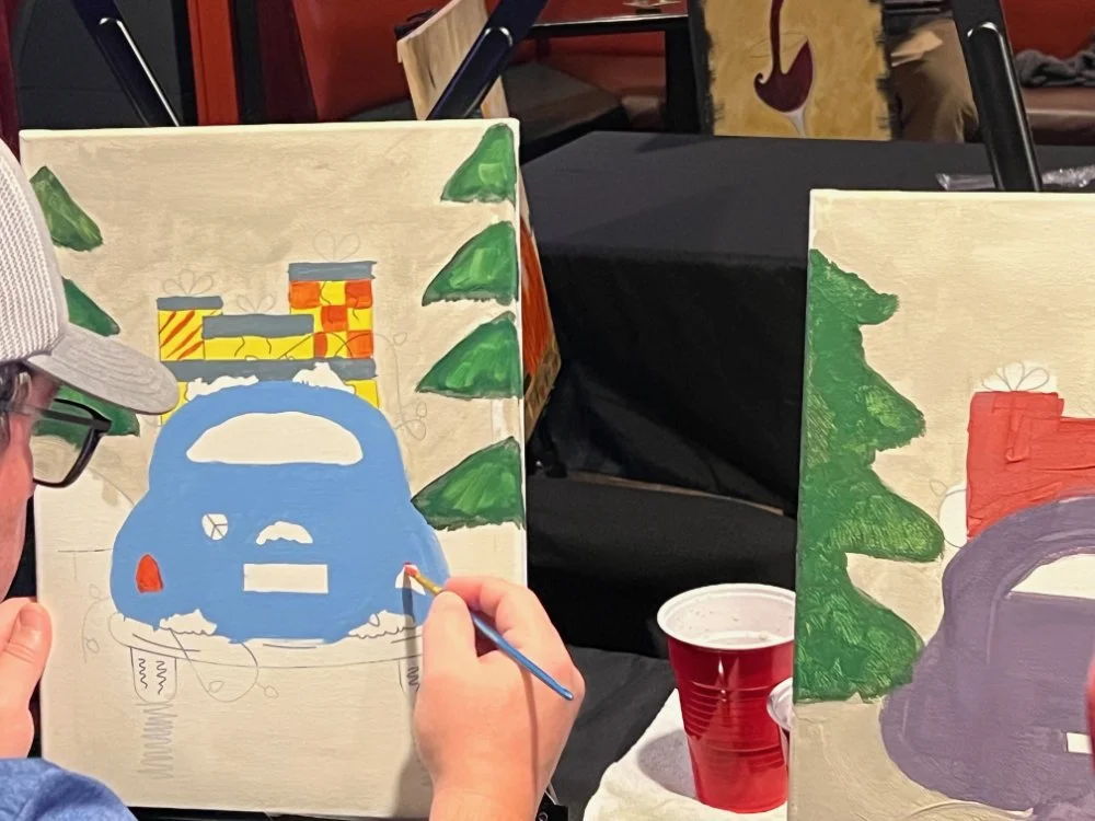 Person painting a Christmas tree on a canvas with a blue train and presents at the top, in a holiday-themed setting.