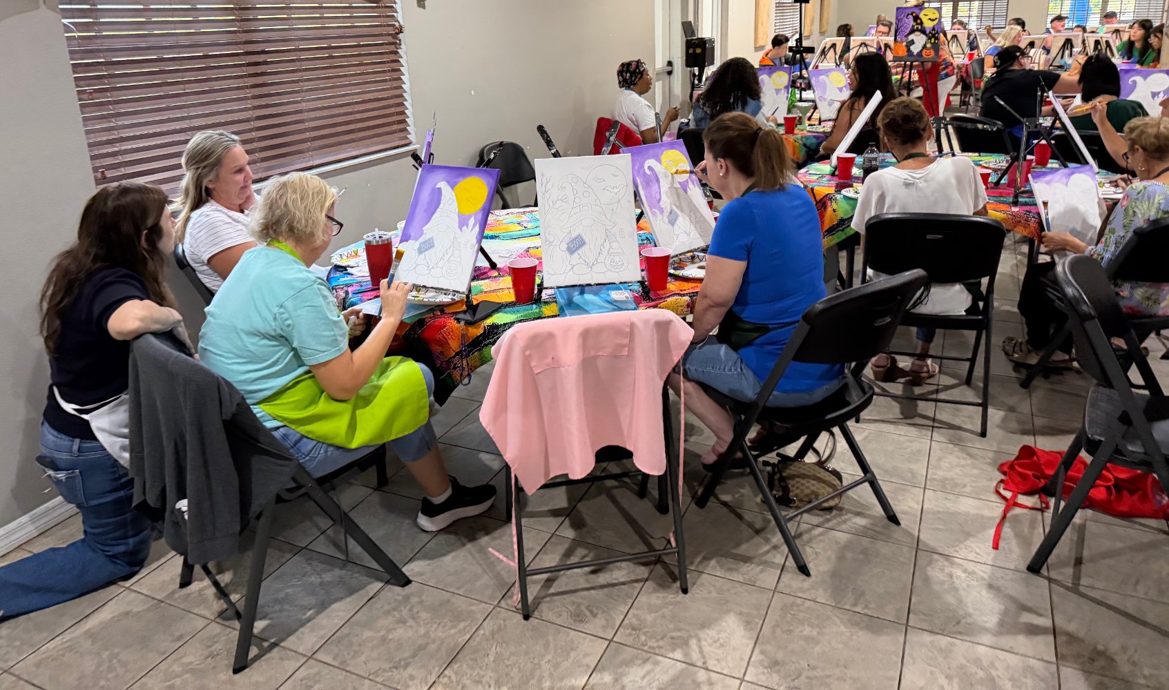 Group of people painting Halloween-themed pictures at a table in a classroom or art studio.