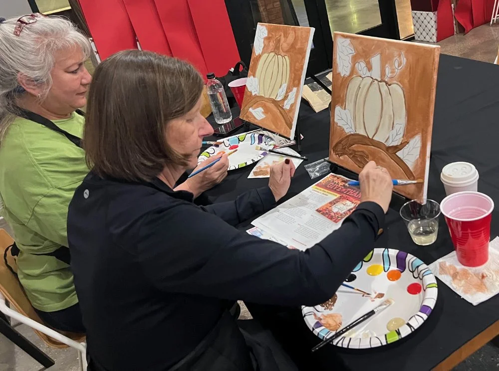 Two women painting pumpkin-themed pictures on canvas at a table during an art class.