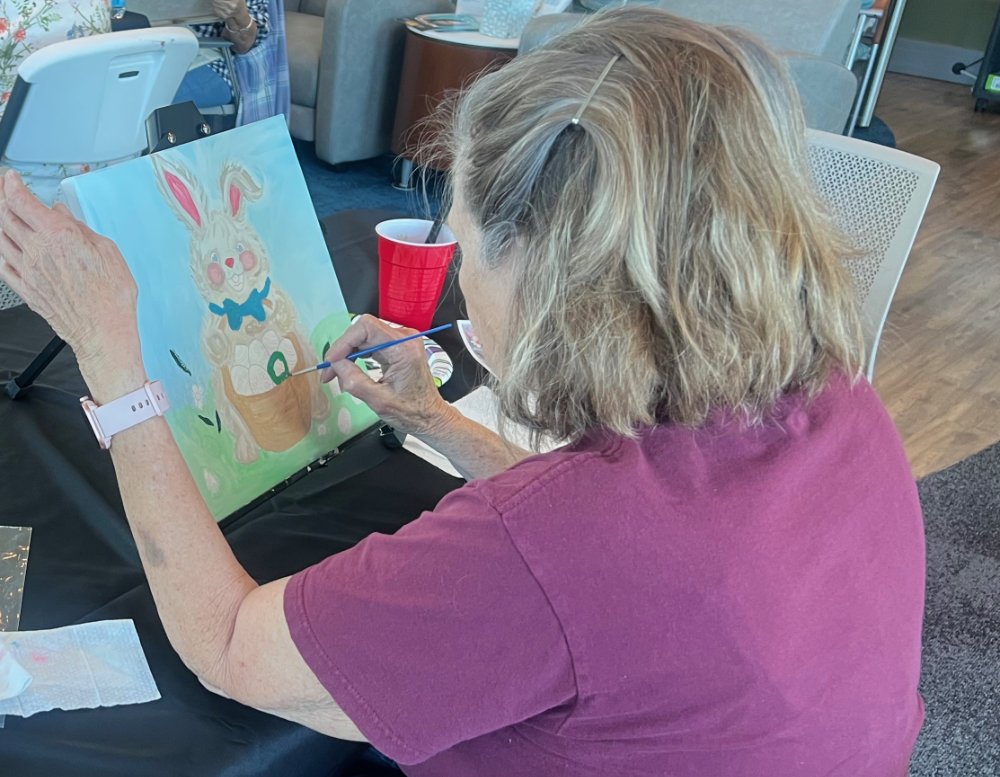 A woman with gray hair and glasses painting an illustration of a rabbit with a blue bow on a canvas at a table.