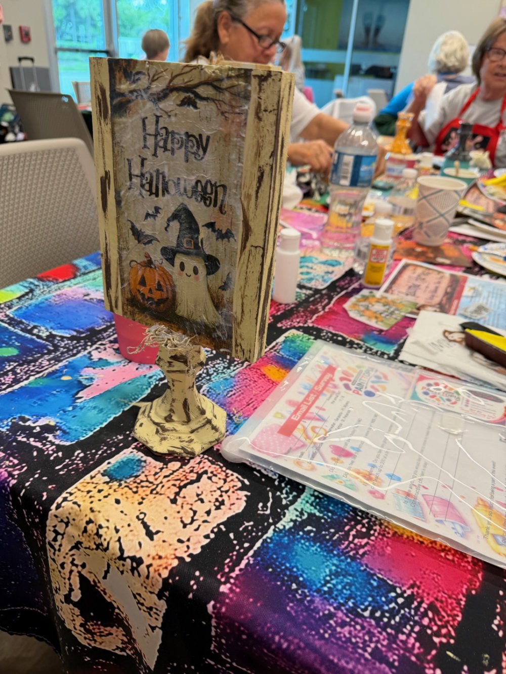 Decorative Halloween-themed sign on a table, with tables, craft supplies, and people in the background.