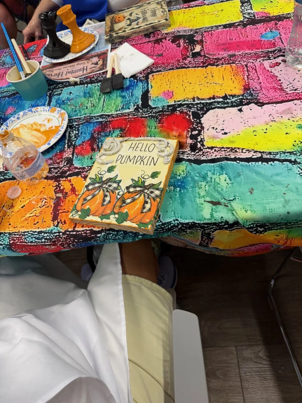 Colorful tablecloth with a hand-painted sign reading 'HELLO PUMPKIN' and decorated with pumpkins and leaves. The table has art supplies, a partially eaten plate of food, a water bottle, and small toys. The scene suggests an arts and crafts activity c