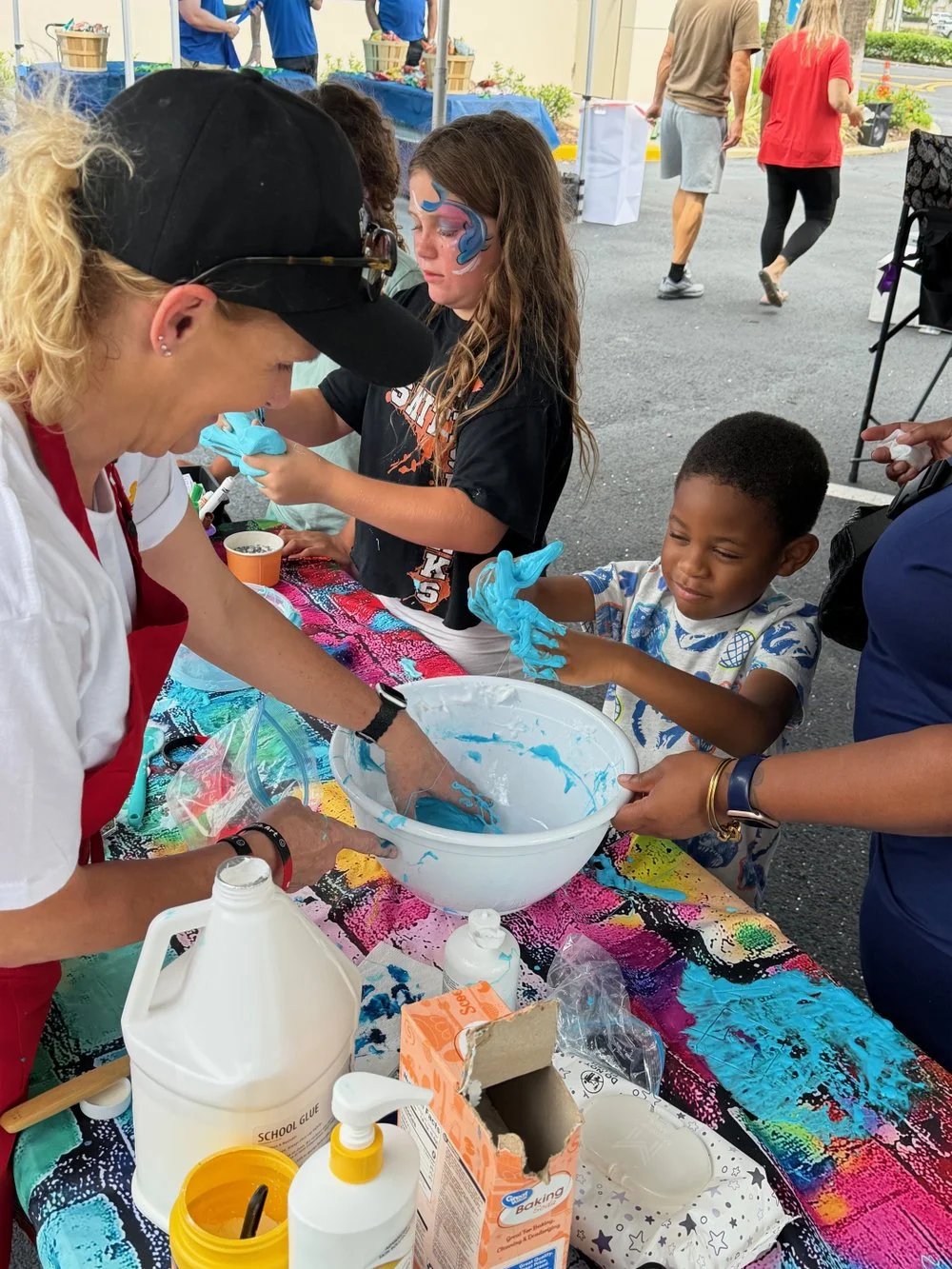 A woman with blonde curly hair, wearing a black cap and red apron, helps children make slime at an outdoor event. A girl with face paint and a boy with short hair are playing with blue slime at a colorful table filled with supplies like glue and baki