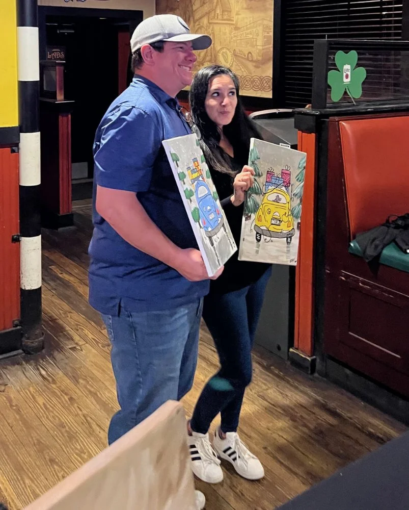 A man and a woman standing inside a restaurant, each holding a colorful painting of a car with gifts on top. The man is wearing a gray cap, blue shirt, and jeans, smiling. The woman has long dark hair, a black top, and is holding her painting with a 
