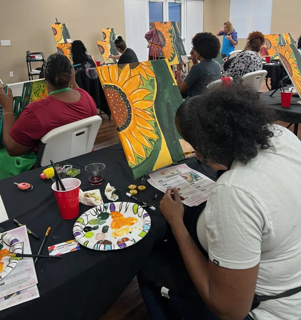 People participating in a group painting class, creating sunflower artwork on canvases, with painting supplies on the tables.