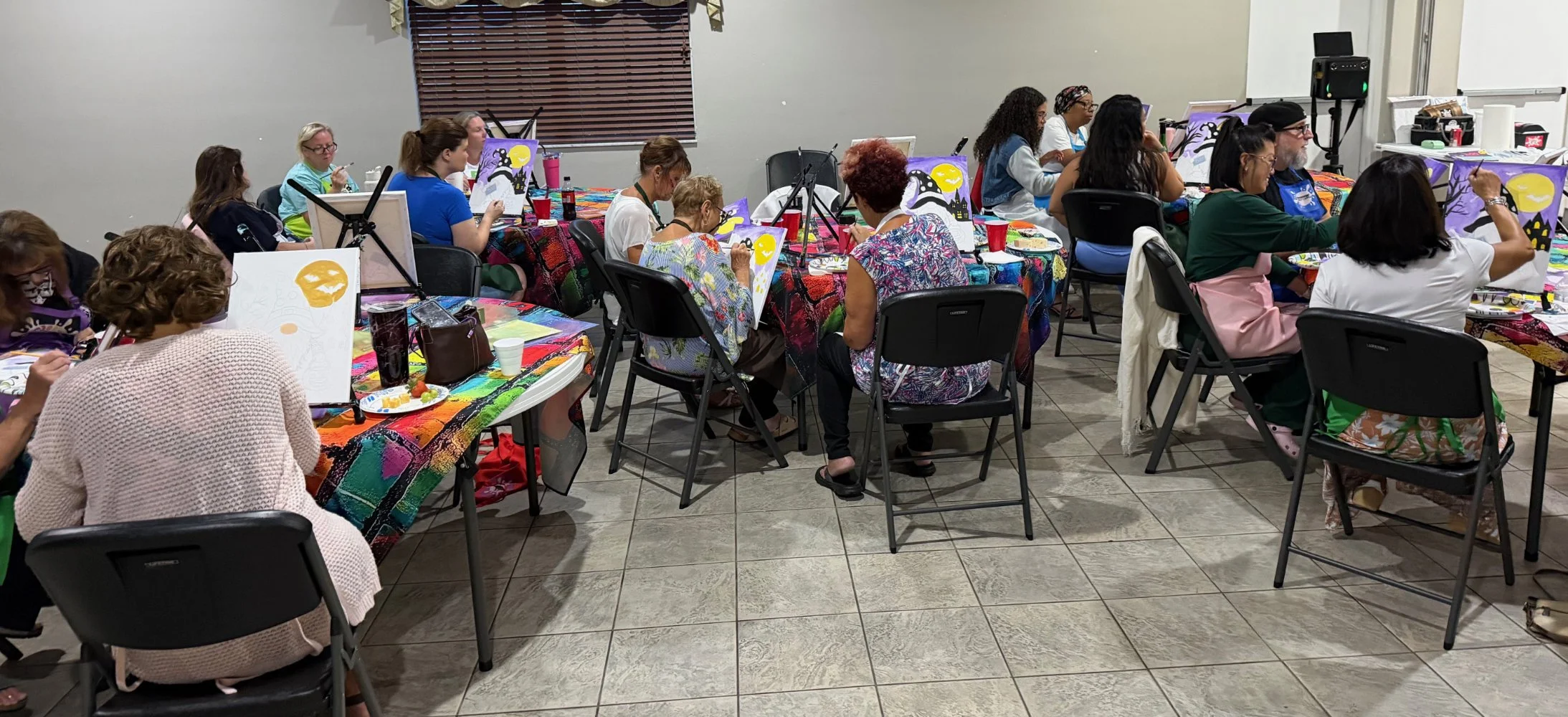 People seated at tables painting Halloween-themed artwork in a classroom or workshop setting.