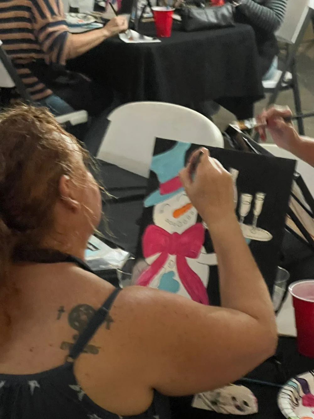 Woman painting a snowman with a pink bow, wearing a blue hat and scarf, at a crowded indoor event.