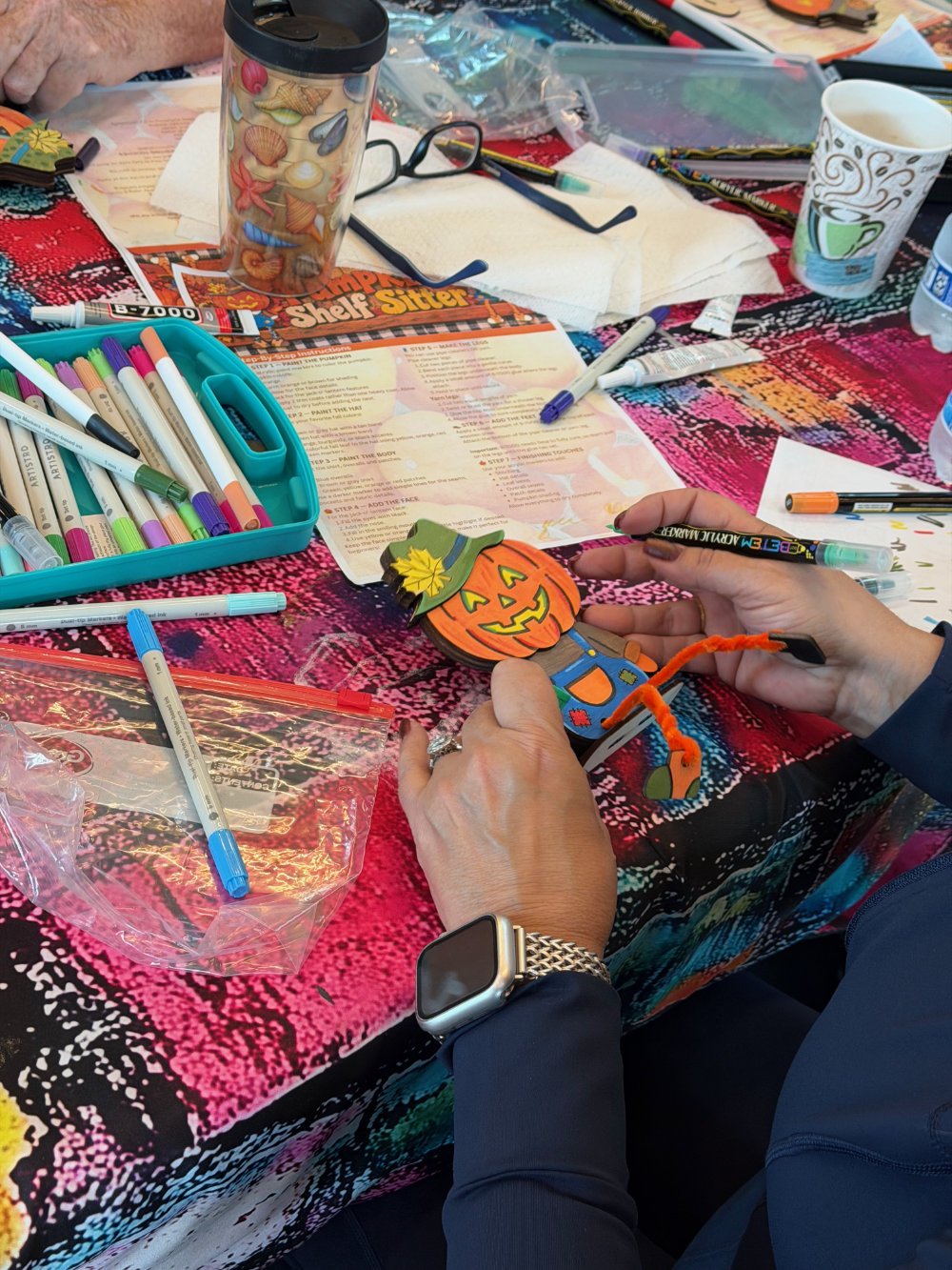 Person holding a colorful pumpkin craft with black markers on a craft table filled with markers, pens, paper, and Halloween decorations.
