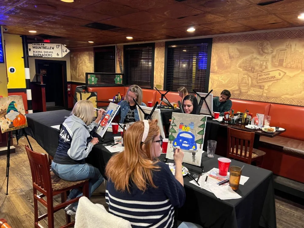 People painting on canvases at a painting class in a cozy restaurant or bar, with drinks and snacks on the tables.