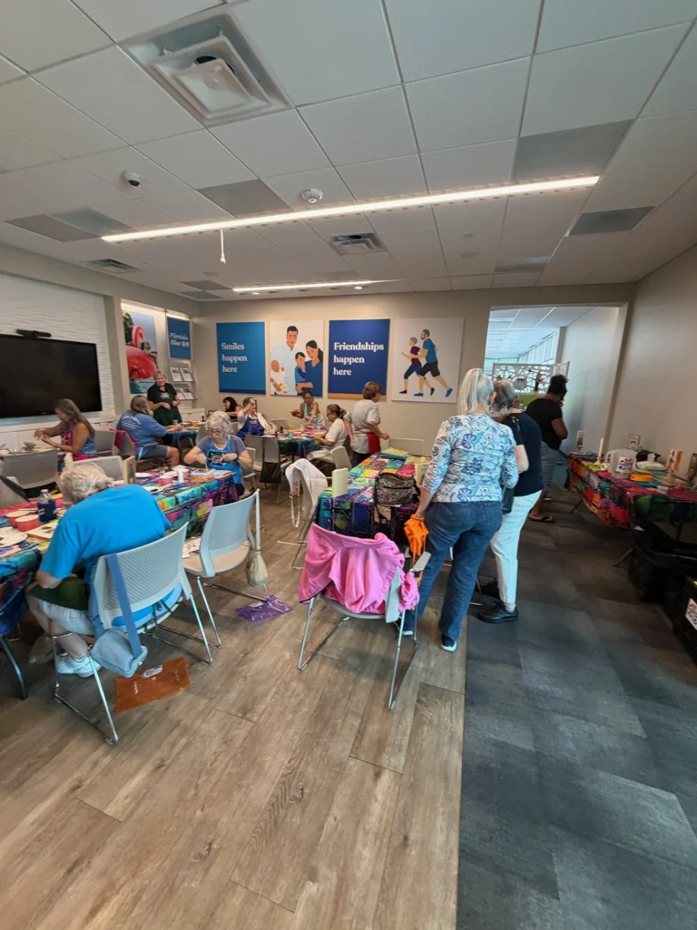 A group of mostly elderly women socializing in a community room with tables covered in colorful tablecloths, some seated and some standing. There are posters on the wall that say 'Smiles happen here' and 'Friendships happen here.'