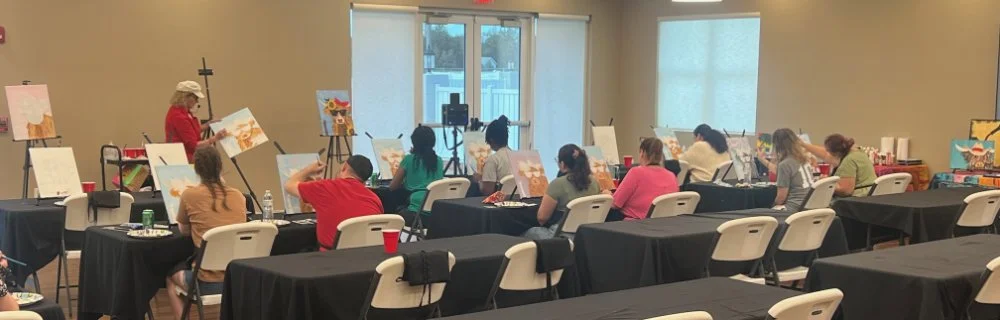 Group painting class with students seated at tables, painting on canvases, instructor standing at the front demonstrating, in a well-lit room with large windows.