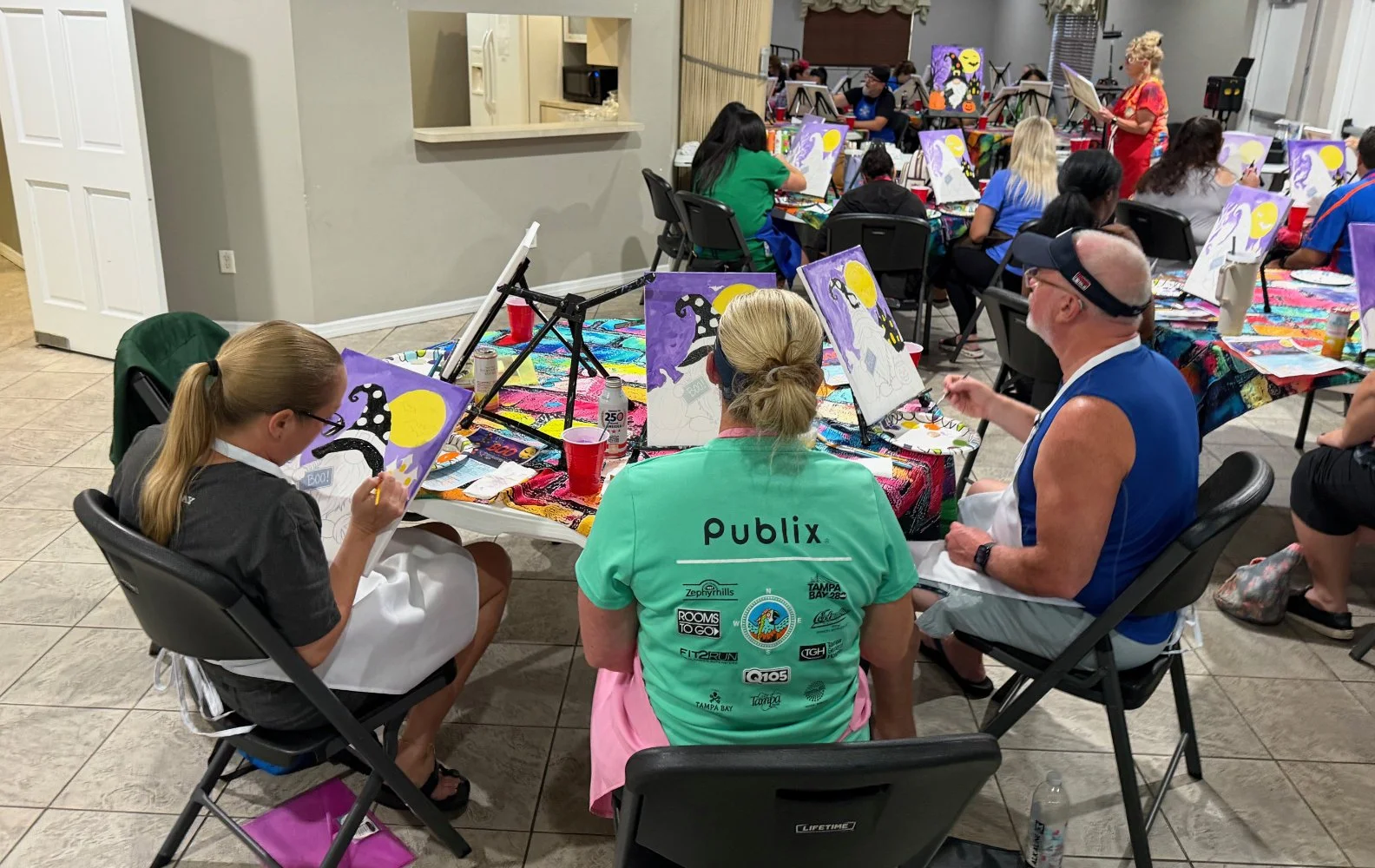People participating in a group painting class indoors, with several individuals working on Halloween-themed canvases depicting black cats, pumpkins, and a moonlit night sky. Participants are seated at tables covered with colorful tablecloths, with a