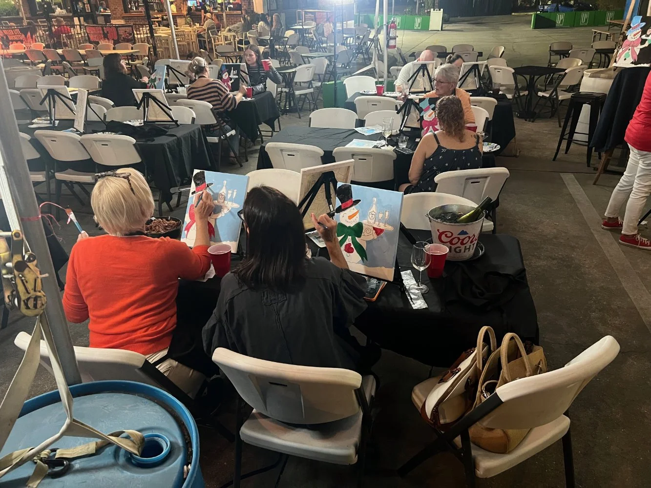 People sitting at tables painting holiday-themed artwork, with an outdoor parking lot in the background and a Coors Light bucket on the table.