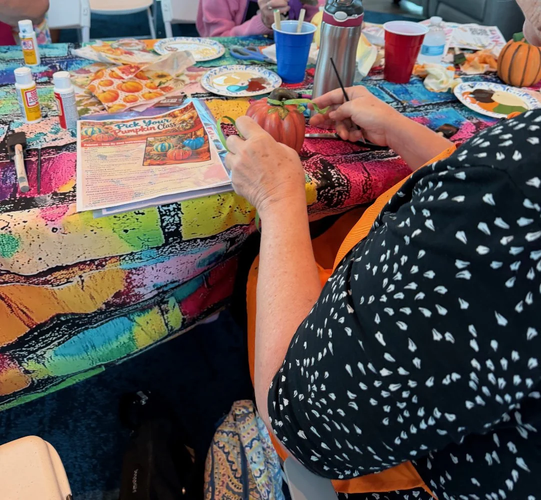 A person painting a pumpkin at a craft table, with various art supplies and colorful decorations visible on the table.