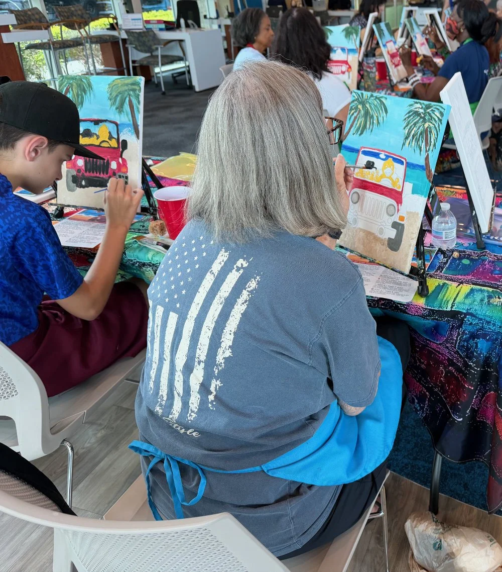 People sitting at tables painting canvas pictures of a Jeep with palm trees and tropical scenery, participating in a group art activity.