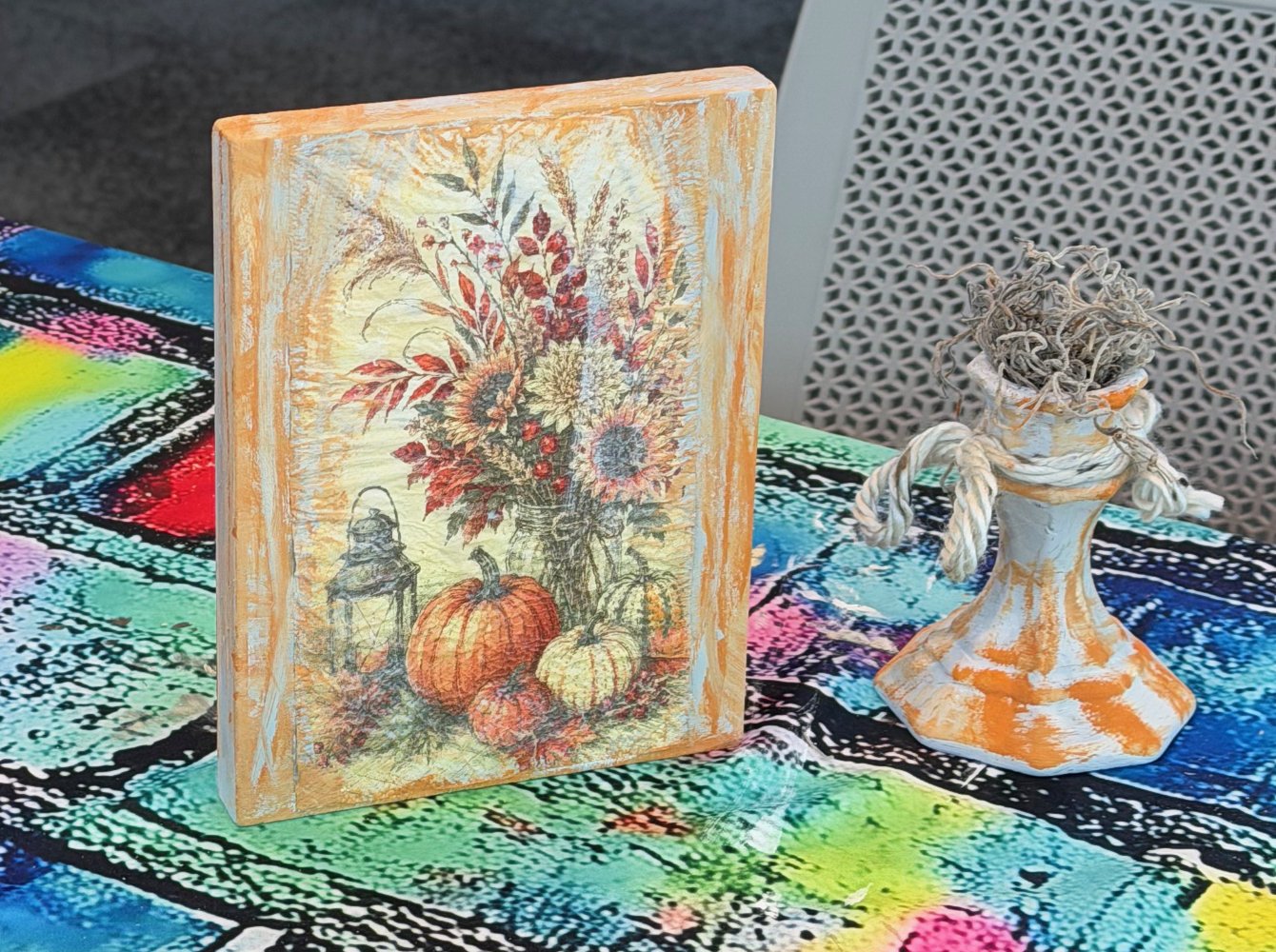 Decorative art piece featuring a pumpkin and sunflower centerpiece on a canvas and a ceramic vase with dried twigs on a colorful tablecloth.