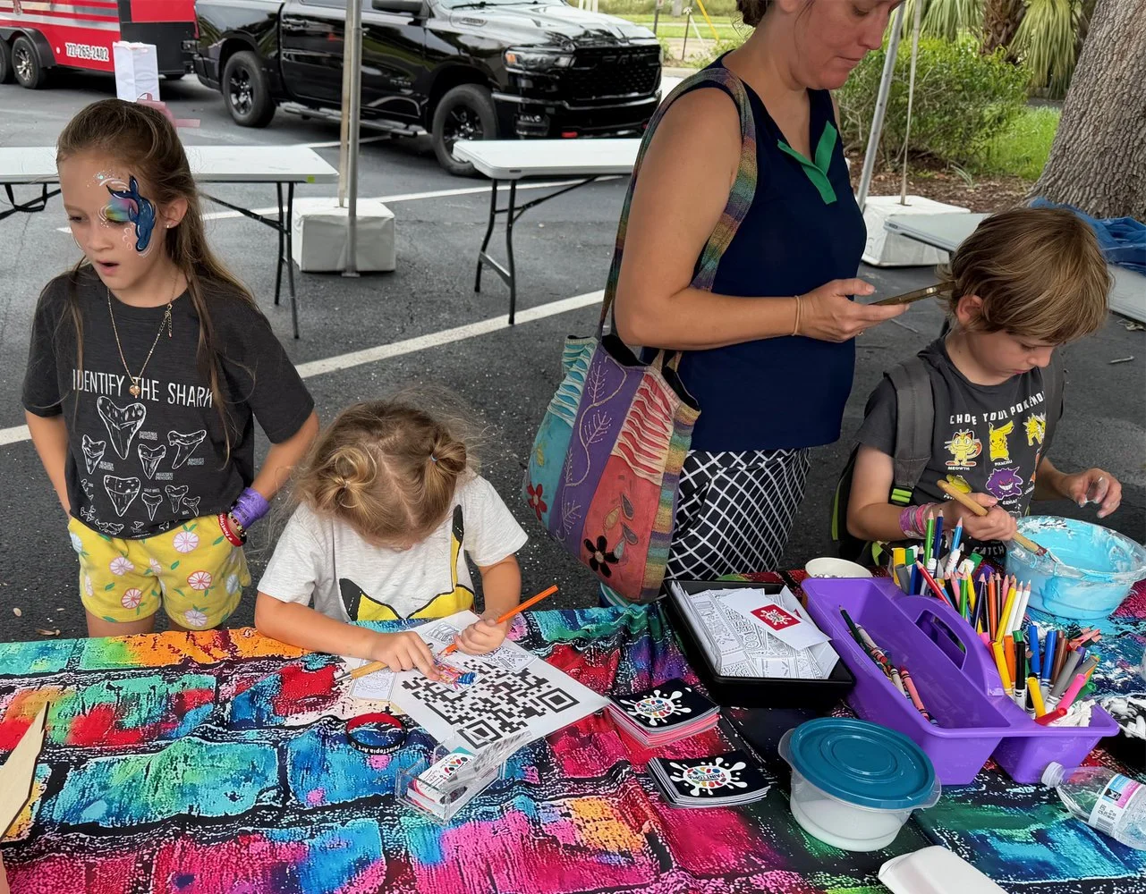 Three children and an adult at an outdoor art and craft booth. One girl has face paint of a blue dolphin, while the other two children are drawing or crafting at the table with colorful supplies.
