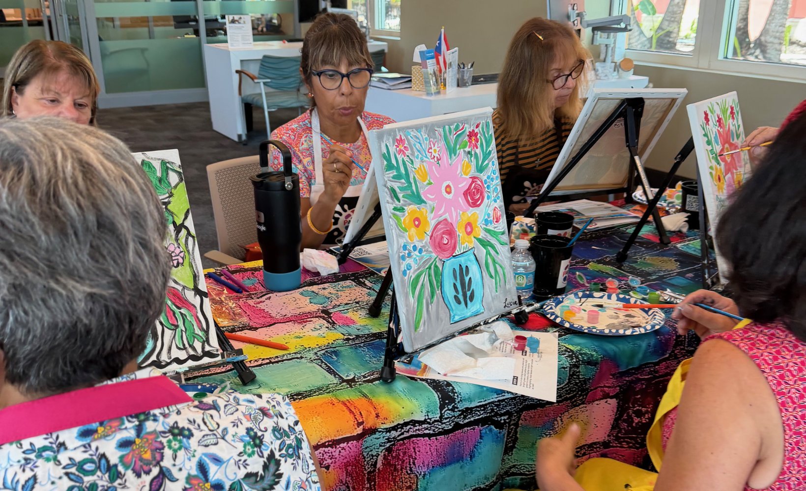 A group of women participating in a paint and sip art class, creating colorful floral paintings on canvases set on small easels.