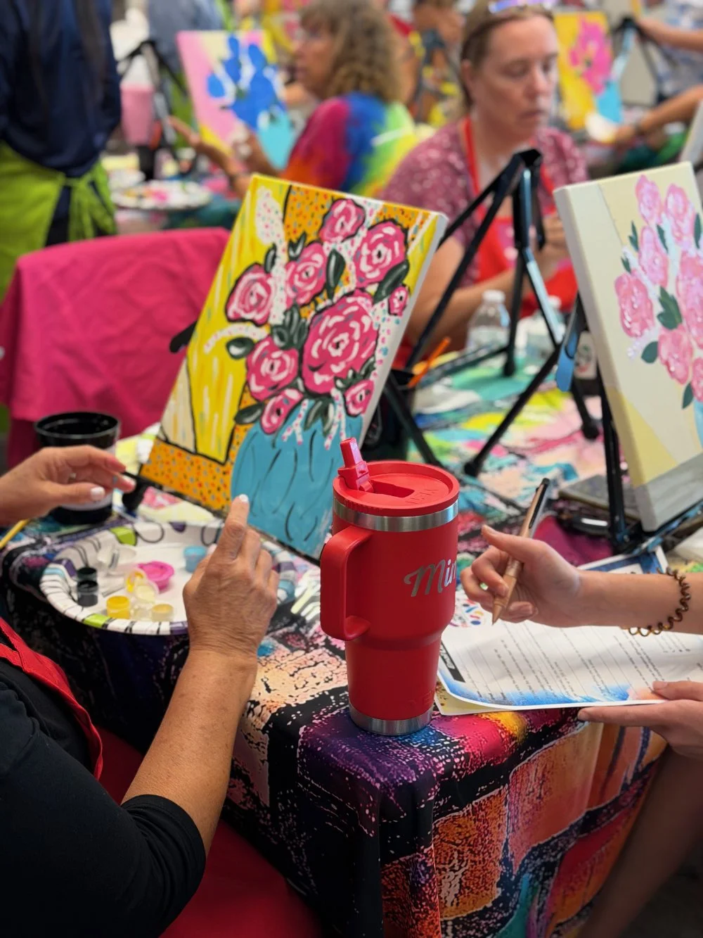 People participating in an acrylic painting class, creating colorful floral artworks on canvases. One person points at a painting of pink roses in a blue vase, with painting supplies and a red insulated mug on the table.