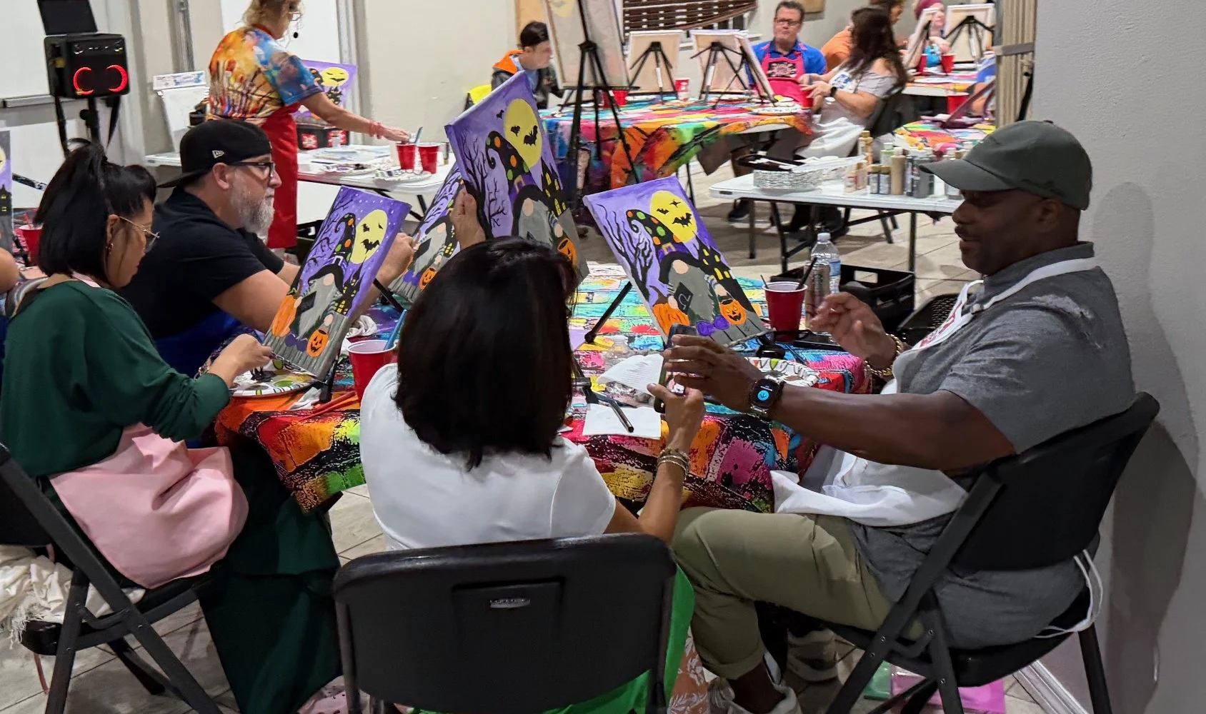 A group of people participating in a Halloween-themed painting class, with some creating spooky scenes featuring a haunted house, pumpkins, and bats on canvas, while others observe or converse, in a room with tables covered in colorful tablecloths an