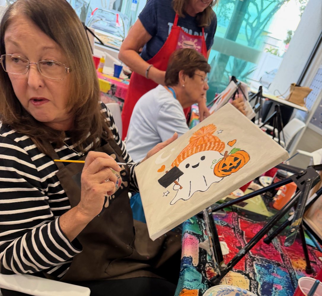 Woman painting a Halloween-themed picture of a friendly ghost holding a marshmallow and a pumpkin with a carved face, wearing an orange striped hat, in a craft workshop.