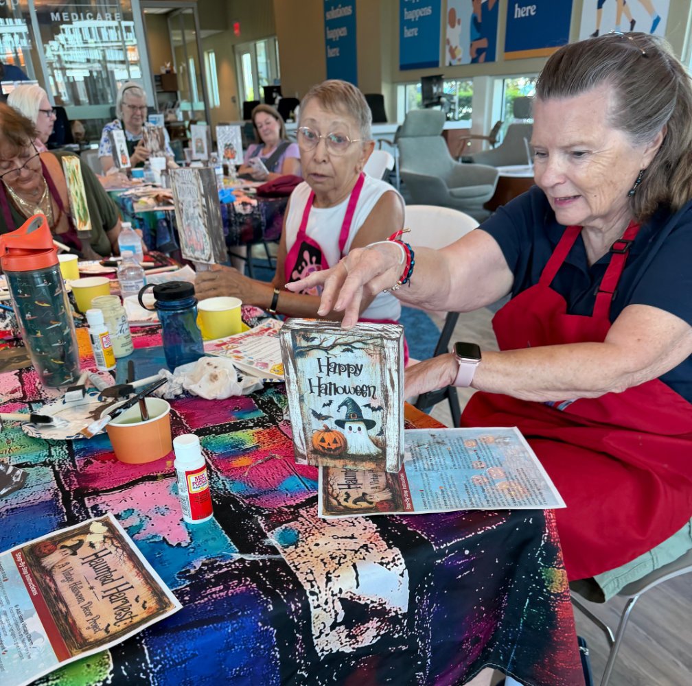 A group of women sitting at a table participating in a craft activity, with a Halloween theme. One woman in the foreground holds a small sign that says "Happy Halloween" with a pumpkin, ghost, and bat illustration. The table is covered with colorful 