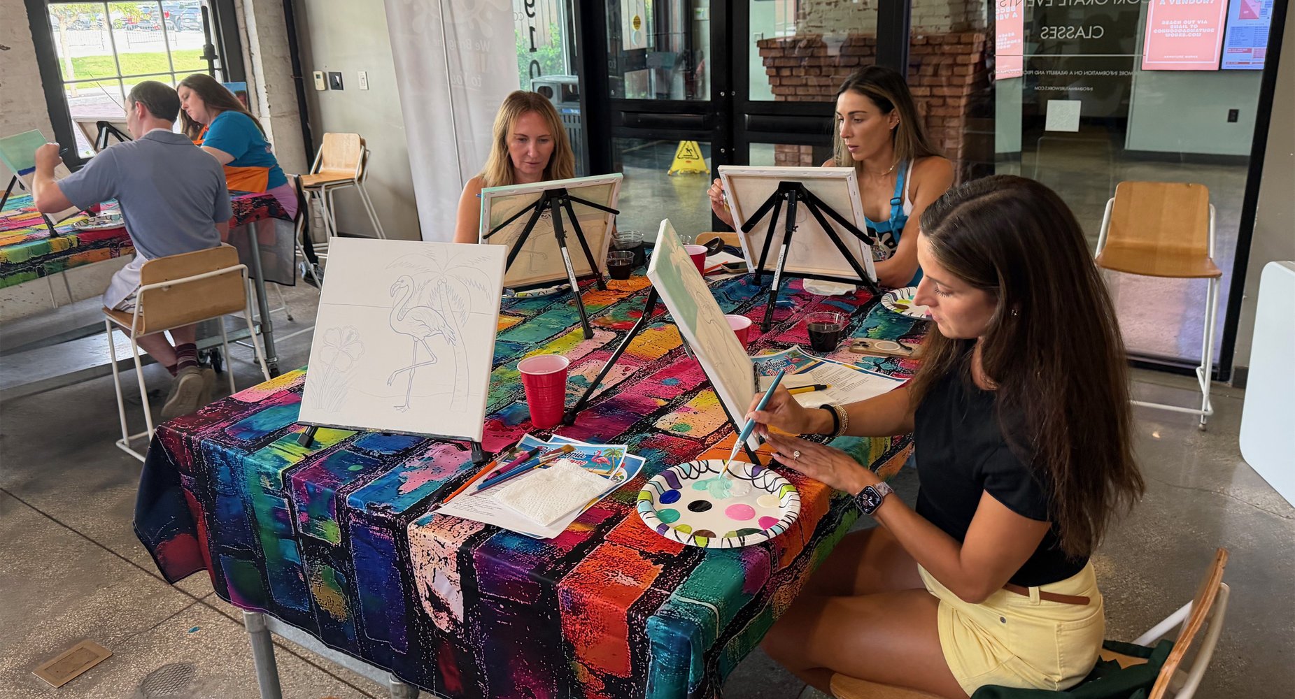 Women painting at a table with a colorful tablecloth in a studio or classroom setting. One woman in yellow shorts is mixing paint on a palette, while others work on canvases placed on easels. There are art supplies and drinks on the table.