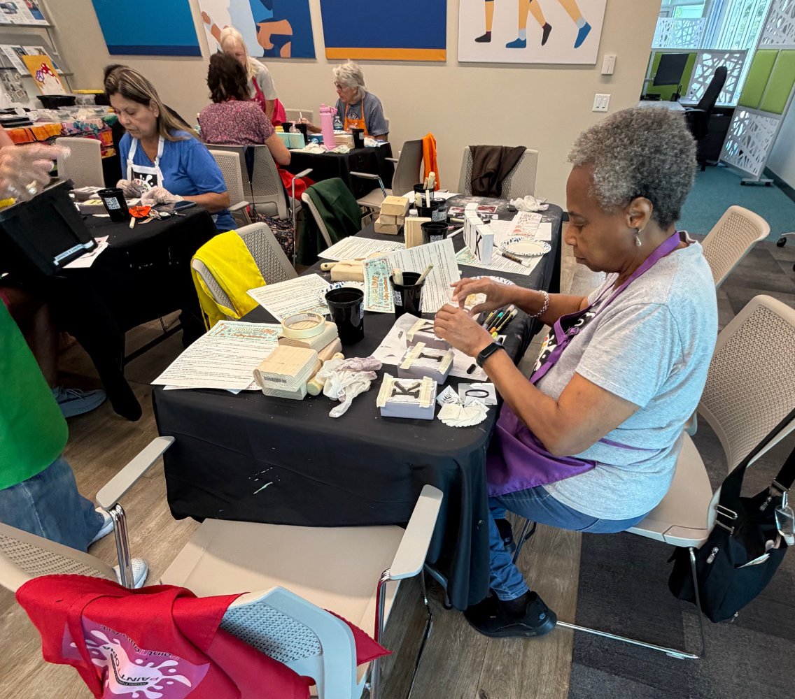 A woman with gray hair working on arts and crafts at a table covered with paper, tape, and boxes, in a room with other women also engaged in similar activities.