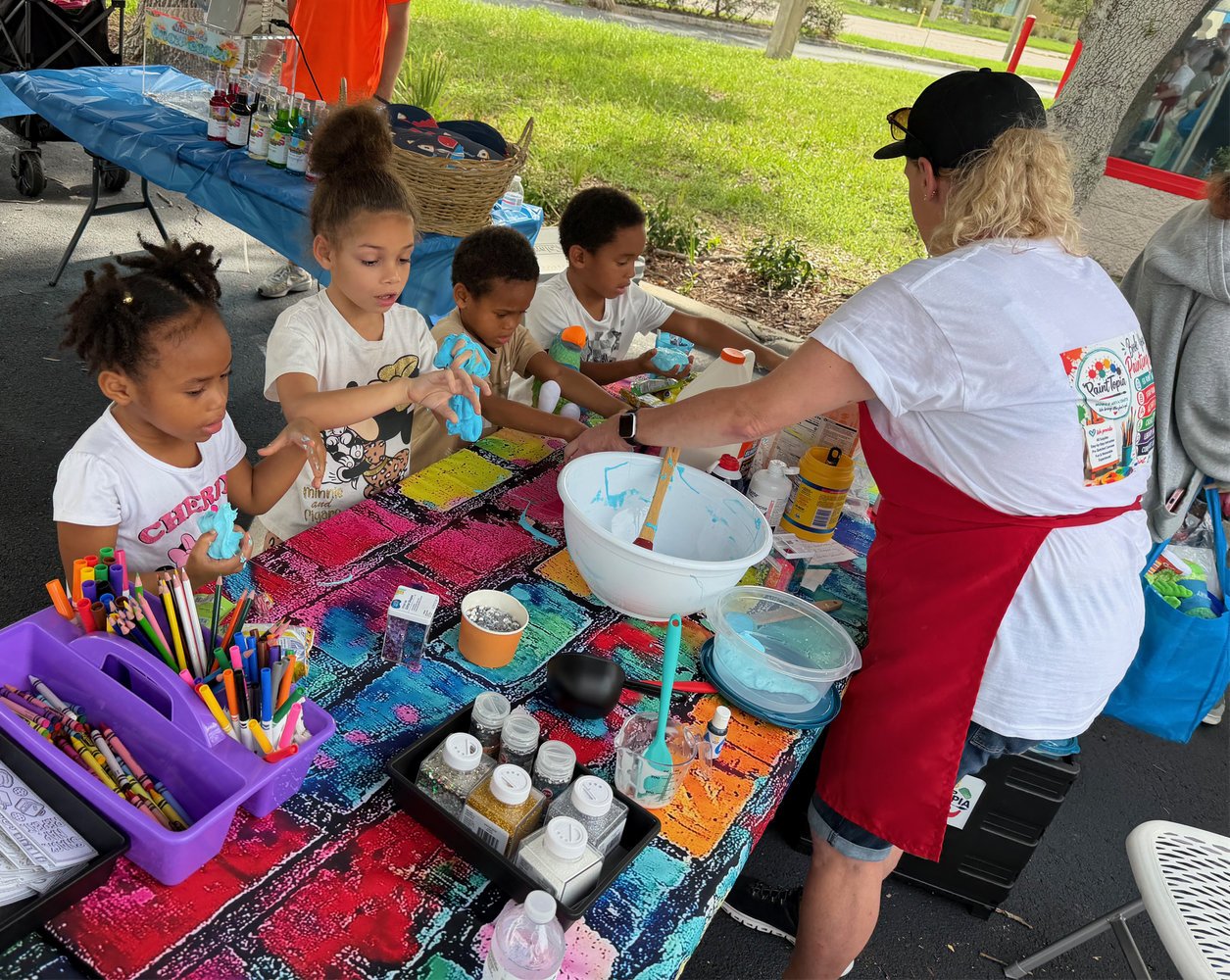 Children participating in a tie-dye workshop at an outdoor event, with a person guiding them, colorful dye supplies, and a vibrant tablecloth.