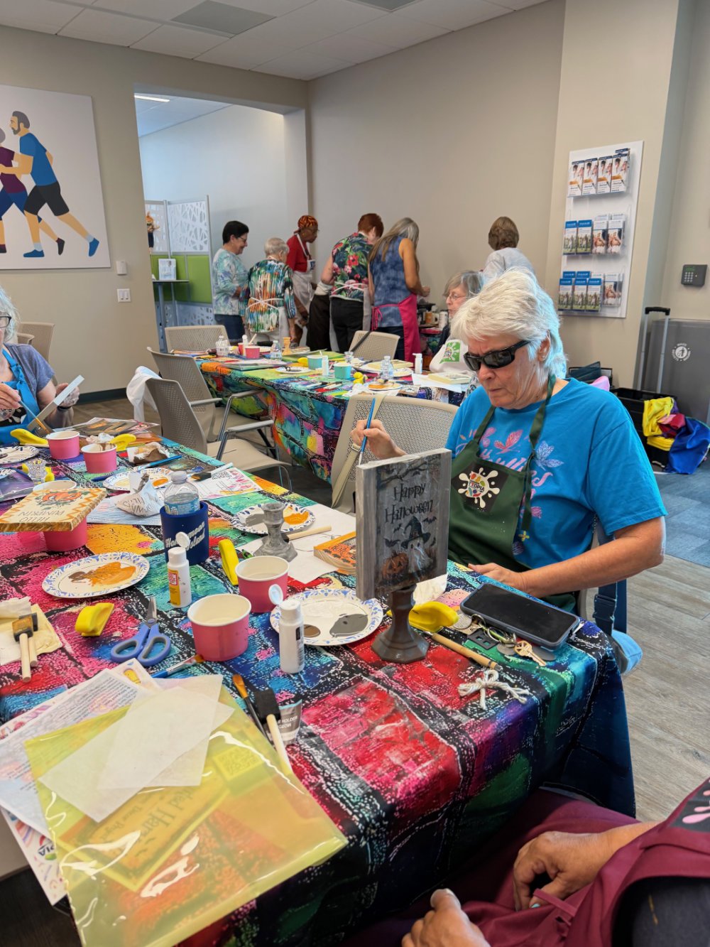 Older women gathered around a table participating in an arts and crafts activity, with art supplies and colorful decorations on the table, in a community room.