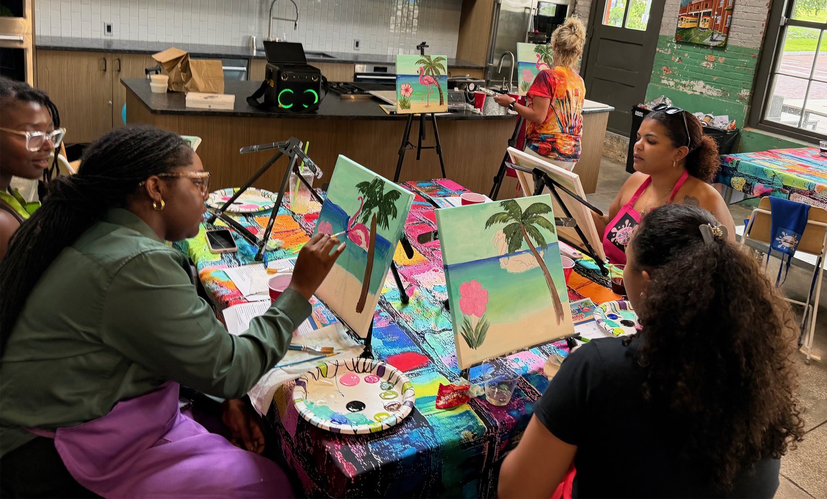People participating in a group painting class in a brightly decorated room, with canvases featuring tropical scenery, and a table covered with a colorful cloth and painting supplies.