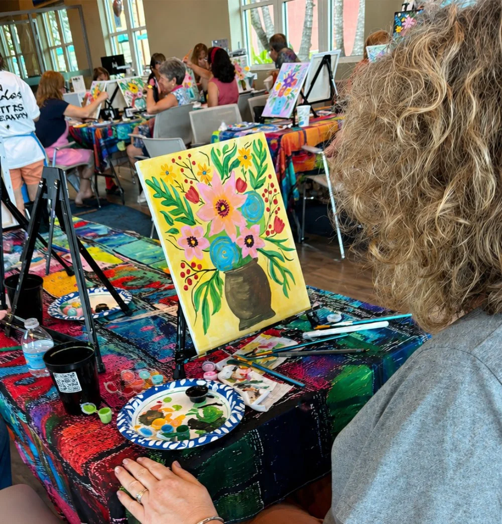 A woman paints a colorful floral still life on canvas at a group art class with other people in the background, working on their own paintings at tables covered with vibrant tablecloths.