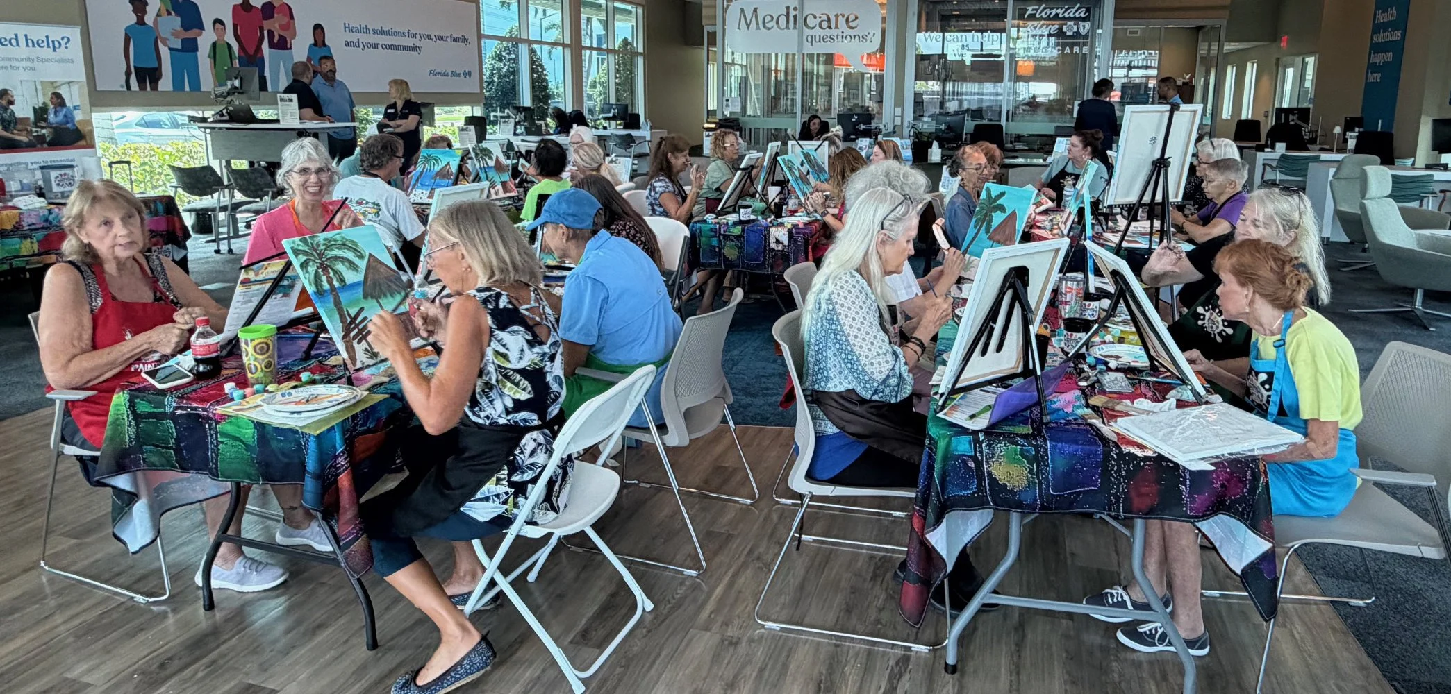 A group of older adults participating in a painting class indoors, seated at tables covered with colorful tablecloths, painting tropical landscape scenes, with large windows and a healthcare signage in the background.