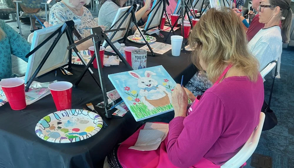 Woman in a pink shirt painting a picture of a white bunny in a basket with flowers and grass at an paint-and-sip event.