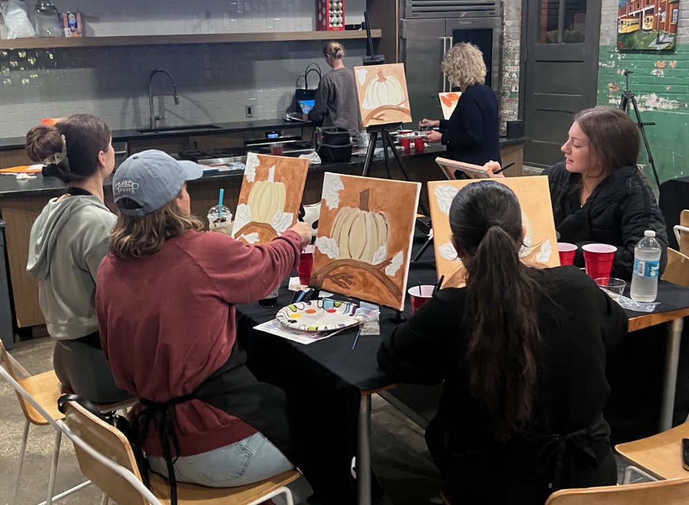 Group of people painting autumn-themed canvases featuring pumpkins and white leaves in an art studio.