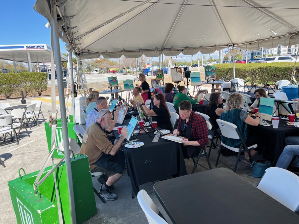 People participating in a painting class under a large white tent outdoors. Students are seated at tables, painting on small canvases, with art supplies and drinks on the tables. Several paintings are displayed on easels in the background.