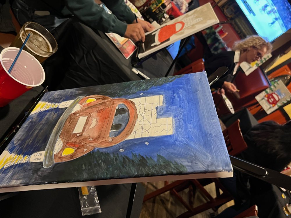 Painting of a red race car on a chocolate brown racing track surrounded by lush green trees, painted on a canvas near people in a meeting room.