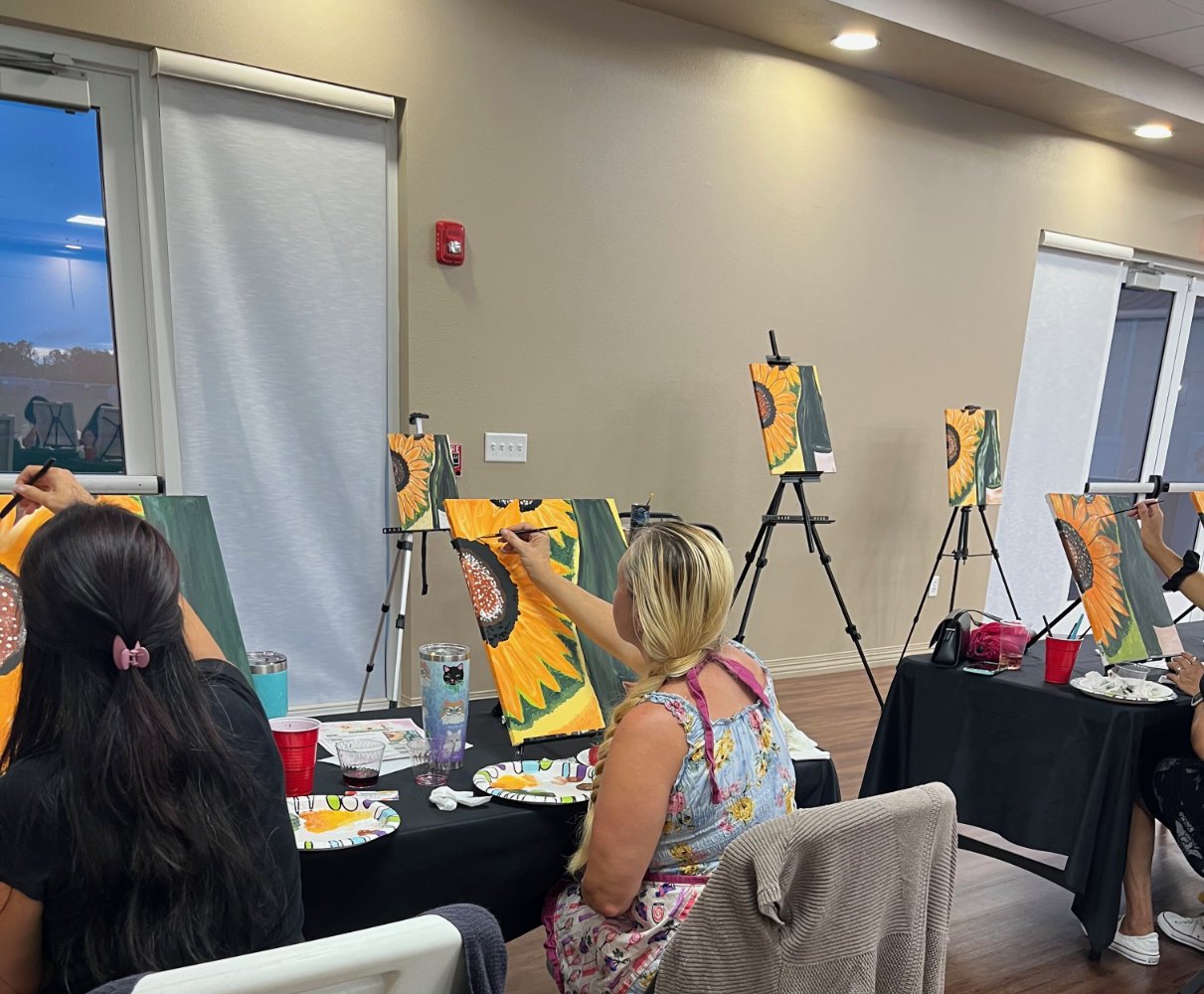 People painting sunflower pictures on canvases at a painting party in a room with large windows and a neutral-colored wall.