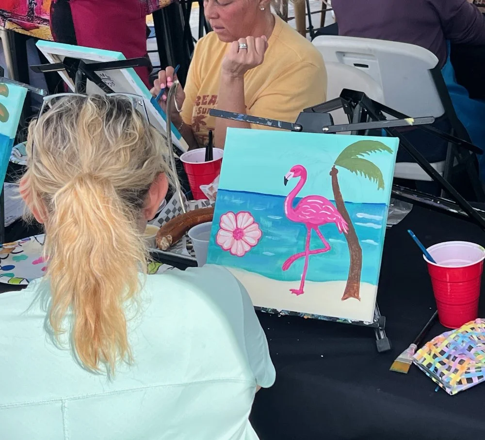 A woman with curly blond hair painting a beach scene with a pink flamingo, a flower, a palm tree, and ocean on a canvas at a social painting event.