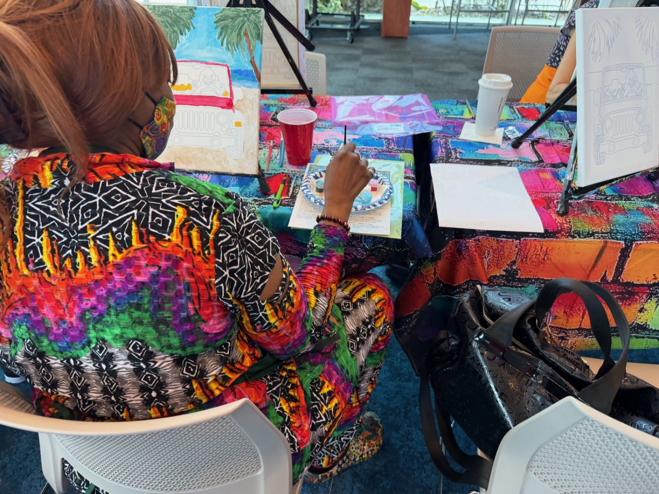 A woman in colorful, patterned clothing and a face mask is painting on a circular palette at a colorful table during an art activity. There are painting supplies, cups, and framed artwork on the table, with a bright, patterned tablecloth and some eas