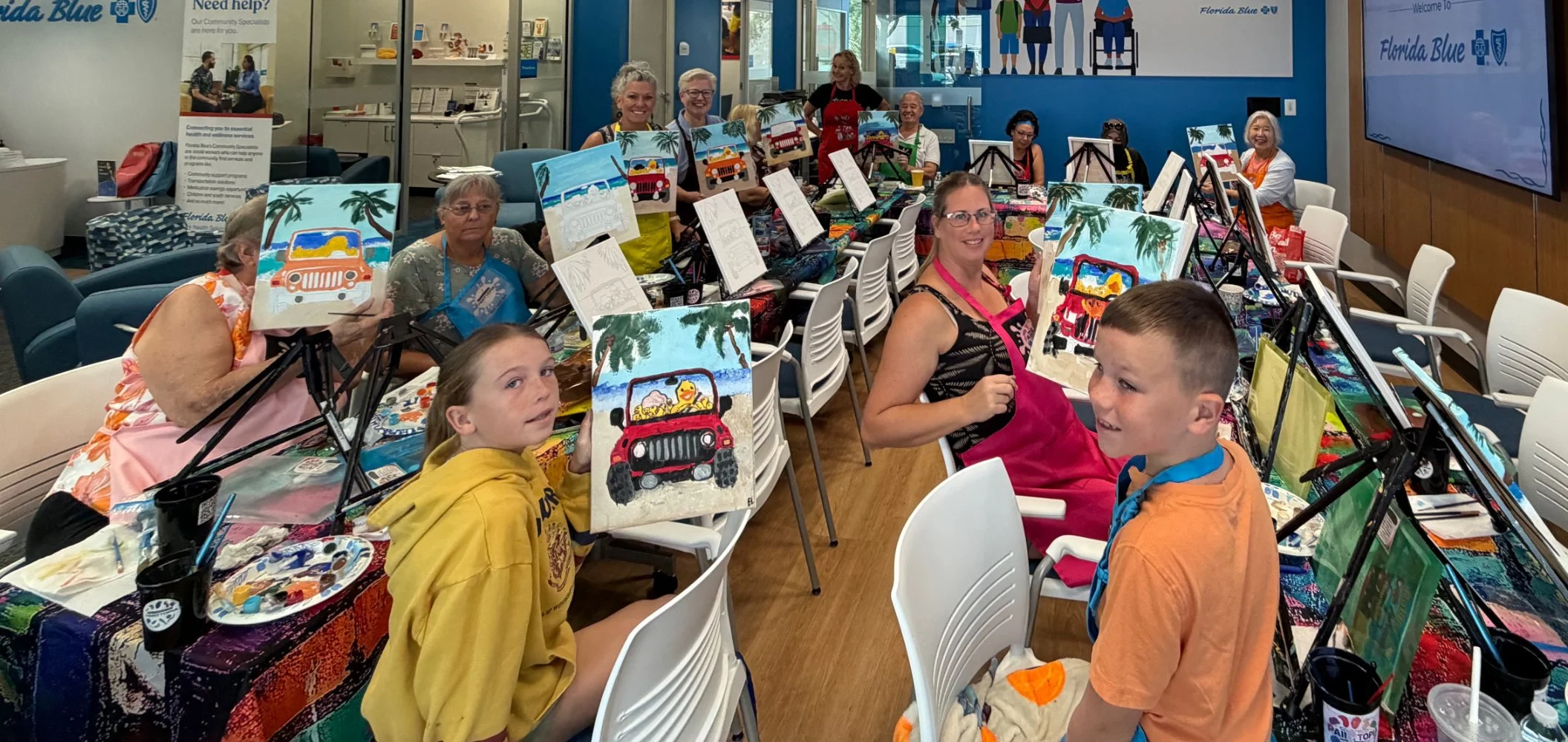 A group of people at a painting class, holding up their colorful beach-themed paintings featuring boats and palm trees, with smiles and art supplies on the table.