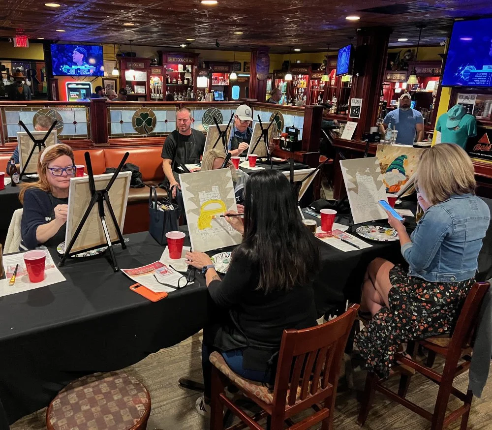 People participating in a group painting activity at a restaurant or bar, with canvas boards, paints, and brushes on the table.