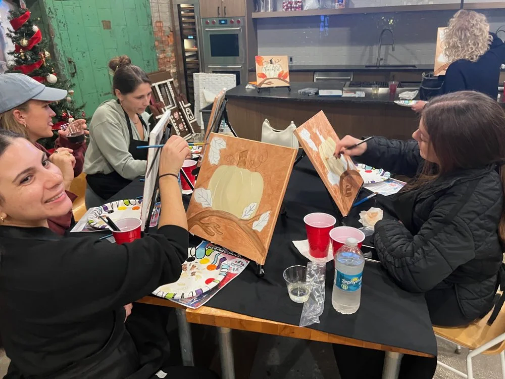 Women painting pumpkins on canvases at an indoor paint-and-sip event.