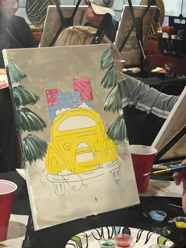 A painting in progress of a yellow vehicle, likely a van, on an easel, with Christmas-themed decorations including green pine branches and pink and blue gift boxes painted on the canvas, in a group setting with other artists and supplies visible.