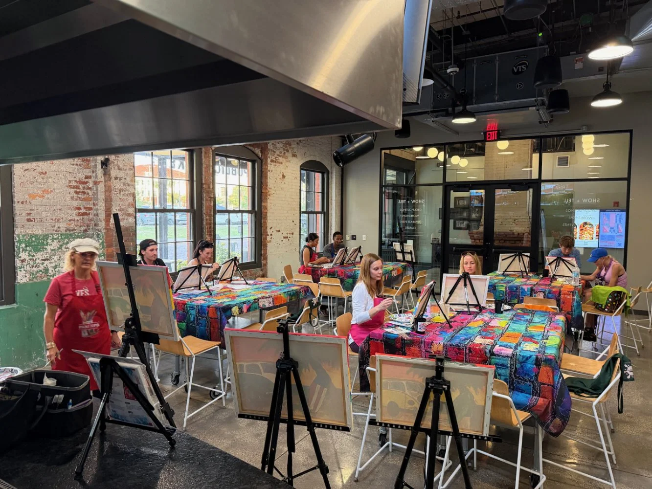 People participating in a group painting class, seated at tables covered with colorful patchwork tablecloths, with easels and canvases in a spacious, industrial-style room with large windows.