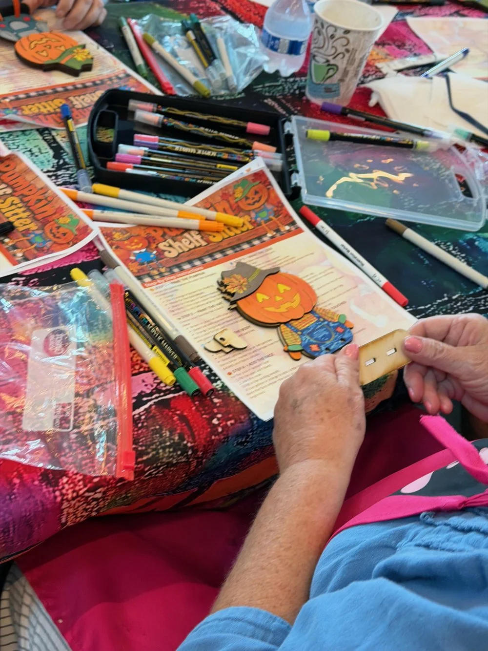 A table with various markers, pens, paper, and Halloween-themed craft decorations, including a pumpkin figure, in a crafting or decorating activity.