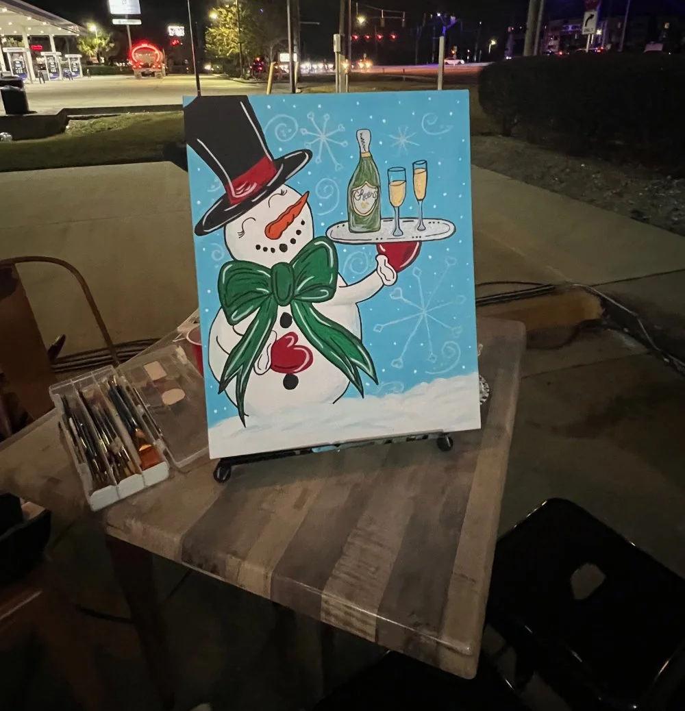 Painted snowman wearing a black top hat with a red band, green scarf with a bow, and orange nose, holding a tray with champagne bottles and glasses, set outdoors at night.