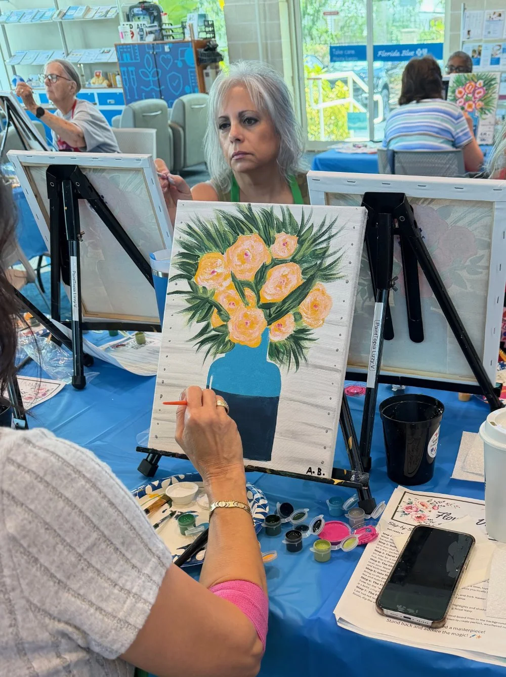 A woman painting a floral picture on a canvas at a paint and sip event. The canvas features yellow flowers in a blue vase with green leaves. Other people are also painting at nearby tables, and art supplies are scattered on the table.