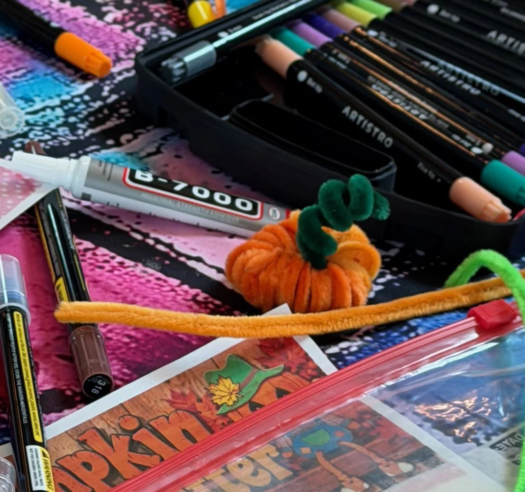 A collection of colorful markers, pens, a white glue bottle, pipe cleaners, and a small orange pumpkin plush toy with a green pipe cleaner stem, arranged on a vibrant pink, black, and white patterned surface.